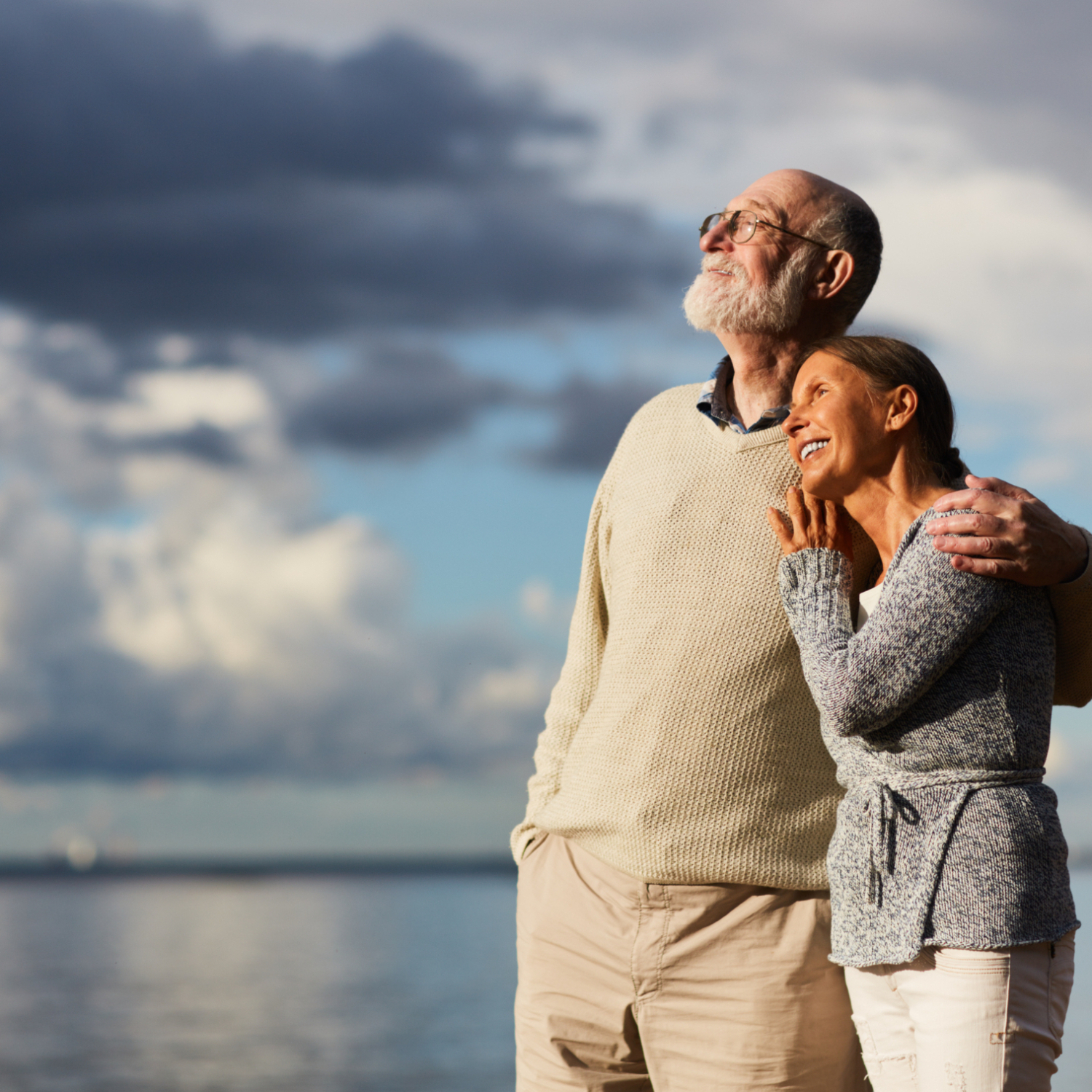 Amorous seniors standing by water against stormy sky and enjoying sundown